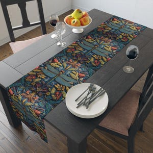 May include: A dark wood dining table is set with a patterned table runner featuring a dragonfly and floral design in teal, orange, and gold. White plates, forks, and wine glasses are arranged on the table, with a bowl of apples.