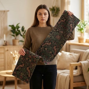 May include: A patterned table runner with a dark base, displaying a repeating design of birds, flowers, and foliage in green, brown, and red. The runner is held up, revealing its length and detailed pattern, suitable for home decoration.