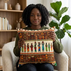 Op de afbeelding: Decoratief kussen met een Afrikaans geïnspireerd ontwerp. Het kussen heeft een crèmekleurige achtergrond met een herhalend patroon van drums, aardewerk en figuren die items op hun hoofd dragen. Het ontwerp bevat tinten bruin, oranje, groen en blauw.