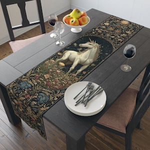 May include: A dark table runner featuring a white unicorn in a floral setting. The runner is decorated with a detailed floral pattern in shades of blue, green, and gold. The table is set with plates, forks, wine glasses, and a bowl of fruit.