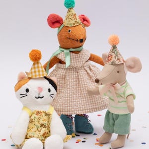 Könnte beinhalten: Drei Plüschtiere mit Partyhüten: eine Katze mit orangefarbenem, weißem und schwarzem Fell, eine braune Maus mit einem grün-weiß gestreiften Hemd und eine orangefarbene Maus mit einem karierten Kleid. Die Spielzeuge sind von buntem Konfetti umgeben.