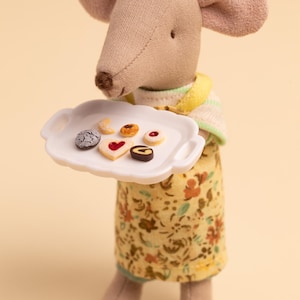 Könnte beinhalten: Eine Stoffmaus-Puppe, die ein gelbes Blumenkleid und ein gestreiftes Oberteil trägt und ein weißes Tablett mit sechs kleinen Keksen hält. Die Kekse haben verschiedene Formen und Verzierungen, darunter ein Herz und Streusel.