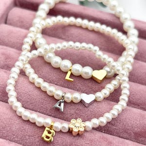 May include: Three pearl bead bracelets with gold and silver charms. The charms include the letters "L", "A", and "B", a gold heart, a silver heart, and a gold flower. The bracelets are displayed on a pink velvet surface.