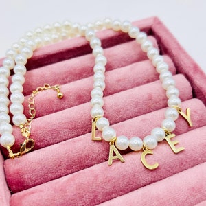 May include: A white pearl necklace with gold letter charms spelling out the name "LACEY". The necklace is displayed in a pink velvet jewellery box.