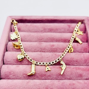 May include: Gold charm bracelet featuring a range of charms, including a horseshoe, a boot, a heart, a star, a gun, and the letter A.