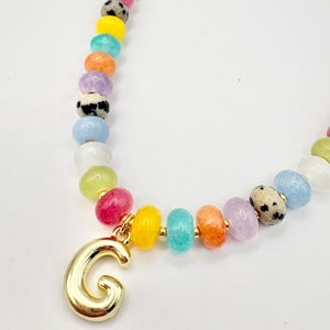 May include: A necklace featuring a variety of colorful, smooth, round beads in shades of yellow, pink, orange, blue, green, white, and black-spotted patterns, accented with small gold-toned spacers and a large, polished gold-toned letter G pendant.
