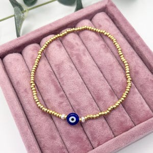 May include: Gold beaded bracelet featuring a blue and white evil eye charm and two small white pearls. The bracelet is presented on a pink velvet jewellery tray. The beads are small and evenly spaced.