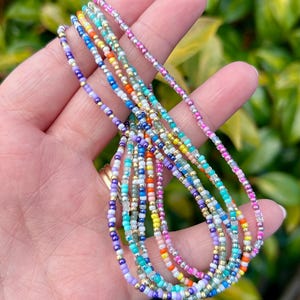 May include: A collection of colorful beaded necklaces in various shades of blue, purple, pink, orange, yellow, and green. The necklaces are delicate and strung with small beads, some with gold accents. The jewelry is held in a hand against a blurred green background.