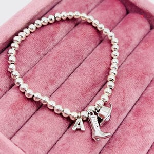 May include: A silver charm bracelet featuring a silver letter A, a silver boot charm, and a silver cowboy hat charm. The bracelet is displayed on a pink velvet background.