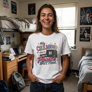 May include: A white short-sleeved t-shirt featuring a colorful graphic print that reads My Childhood Had a Pause Button, accompanied by an illustration of a vintage television set with a pause symbol on the screen and a connected video cassette player.
