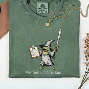 May include: Moss green Comfort Colors t-shirt with a cartoon frog wizard holding a sword and IEP clipboard. The text "We Update IEPs at Dawn" is below the frog. A gold star pendant necklace is draped on the shirt.