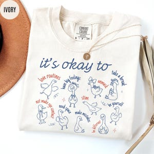 May include: Ivory-colored t-shirt with the phrase "it's okay to" in blue script, followed by various affirmations and goose illustrations. The shirt features a Comfort Colors tag and a gold necklace.