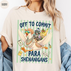 May include: Ivory-colored t-shirt with the text "OFF TO COMMIT PARA SHENANIGANS." The graphic features a goose wearing sunglasses, a raccoon, and a frog riding the goose. The design includes stars and other small graphics.