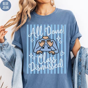 May include: Blue jean-colored t-shirt with the words "All Done!" and "Class Dismissed!" in white script. The design features a cartoon figure with hands up, surrounded by stars and a heart. The shirt has short sleeves.