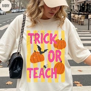 May include: An ivory t-shirt featuring a graphic print with yellow vertical stripes and the text "TRICK OR TEACH" in pink. The design includes three orange pumpkins, a black bat, a black spider, and a black spiderweb illustration.