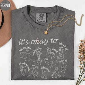 May include: A dark gray Comfort Colors t-shirt with the phrase "it's okay to" and various affirmations in white script and illustrations of ducks. A gold star pendant necklace rests on the shirt.