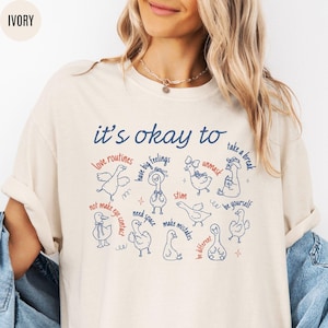 May include: Ivory-colored t-shirt with the text "it's okay to" above a series of illustrated ducks with phrases like "love routines" and "take a break." The design is in navy blue and red.