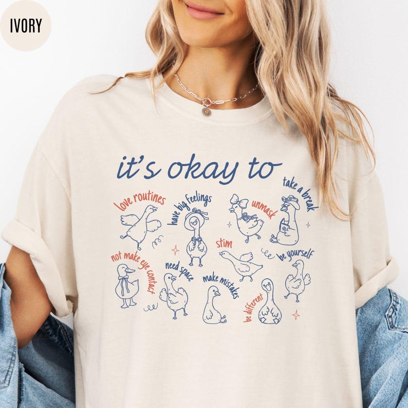 May include: Ivory-colored t-shirt with the text "it's okay to" above a series of illustrated ducks with phrases like "love routines" and "take a break." The design is in navy blue and red.