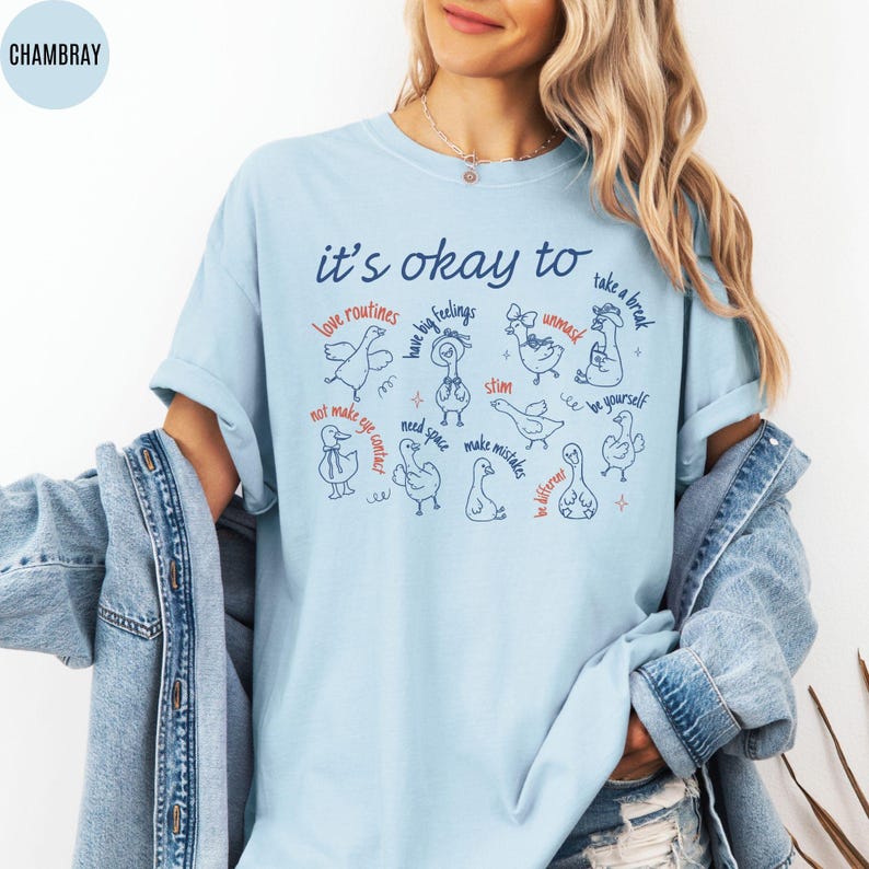 May include: Light blue t-shirt with the text "it's okay to" and various phrases and illustrations of ducks. Phrases include "love routines", "have big feelings", and "take a break".