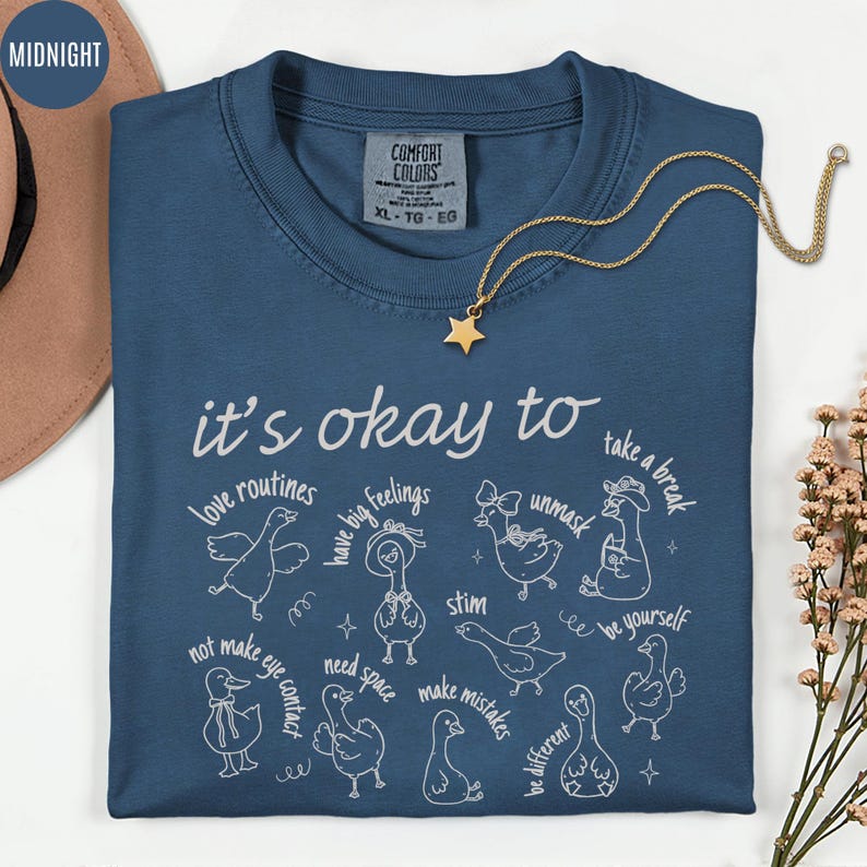 May include: A navy blue Comfort Colors t-shirt with the phrase "it's okay to" and various affirmations, each accompanied by a whimsical duck illustration. A gold star pendant necklace is draped on the shirt.