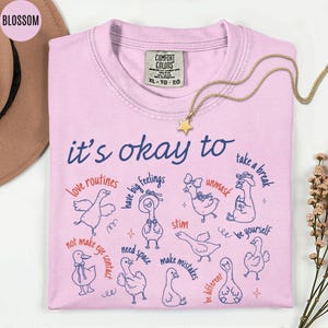 May include: A light pink Comfort Colors t-shirt with the phrase "it's okay to" and various affirmations, along with cartoon geese illustrations. A gold necklace with a star pendant is draped across the shirt.