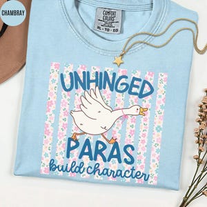 May include: Light blue t-shirt with the words "UNHINGED PARAS build character" and a cartoon goose graphic. The graphic is set against a floral and striped background. A gold star necklace is draped across the shirt.