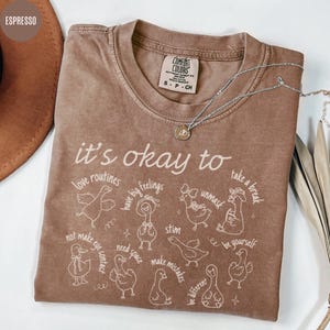 May include: Espresso-colored Comfort Colors t-shirt with the phrase "it's okay to" and various affirmations, along with whimsical duck illustrations. A silver necklace with a pendant is draped across the shirt.