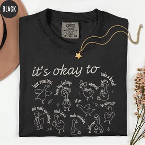 May include: Black Comfort Colors t-shirt with the phrase "it's okay to" and various affirmations, each paired with a whimsical duck illustration. A gold star pendant necklace rests on the shirt.