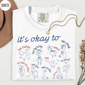May include: White Comfort Colors t-shirt with the text "it's okay to" and various phrases and illustrations of ducks. A gold star pendant necklace is draped across the shirt. The shirt is a size XL.