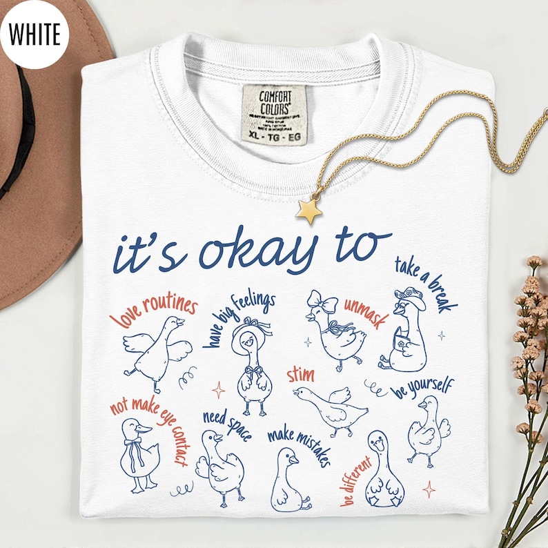 May include: White Comfort Colors t-shirt with the text "it's okay to" and various phrases and illustrations of ducks. A gold star pendant necklace is draped across the shirt. The shirt is a size XL.