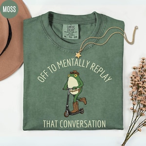 May include: Moss green t-shirt with the text "OFF TO MENTALLY REPLAY THAT CONVERSATION". The shirt features a cartoon frog riding a scooter and wearing a cowboy hat and boots. A gold star pendant necklace is also visible.