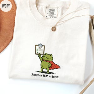May include: Ivory-colored t-shirt featuring a cartoon frog superhero holding a clipboard labeled "IEP." The frog wears a red cape and a green bow tie. The text "Another IEP, m'lord?" is printed below the frog. A gold necklace with a pendant is also visible.