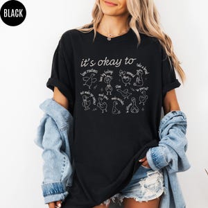 May include: Black t-shirt with white text and illustrations. The phrase "it's okay to" is above a series of duck drawings, each paired with a word or phrase like "love routines" and "be yourself". The word "BLACK" is in a circle in the upper left.