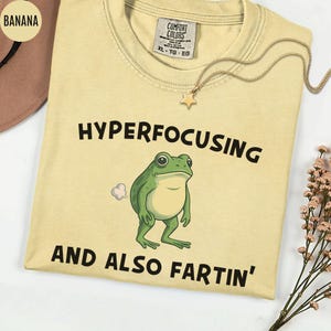 May include: Light yellow t-shirt with the text "HYPERFOCUSING AND ALSO FARTIN'" and a cartoon frog illustration. The shirt has a crew neck and short sleeves. A gold necklace with a star pendant is on the shirt.