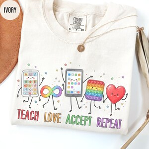 May include: An ivory t-shirt featuring a graphic of five animated characters: a smartphone, an infinity symbol, a tablet, a pop-it toy, and a heart. Below the characters, the text reads "TEACH LOVE ACCEPT REPEAT" in colorful, bold letters.