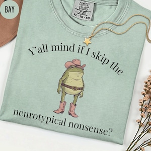 May include: A light green t-shirt featuring a graphic of a frog wearing a pink cowboy hat and pink cowboy boots with a brown belt. The text reads, Y'all mind if I skip the neurotypical nonsense? The shirt has a Comfort Colors label in the neck.