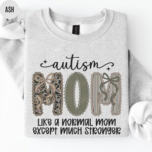 May include: Light gray sweatshirt with the words "autism MOM" in decorative lettering. The word "MOM" is in a camouflage pattern, olive green, and vertical stripes. The text "Like a normal mom except much stronger" is below.