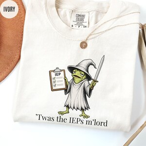 May include: Ivory-colored t-shirt featuring a cartoon frog dressed as a wizard, holding a clipboard labeled "IEP" and a sword. The text "'Twas the IEPs m'lord" is printed below the frog. A gold necklace is draped on the shirt.