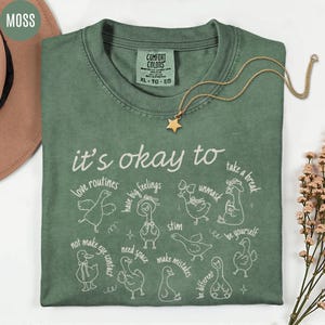 May include: A moss green t-shirt with the phrase "it's okay to" and various affirmations in white script and illustrations. A gold star pendant necklace rests on the shirt. The shirt is labeled XL-TG-EG.