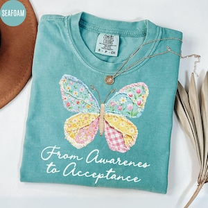May include: Seafoam green t-shirt with a butterfly applique. The butterfly has floral and gingham patterns in pink, yellow, and blue. The text "From Awareness to Acceptance" is below the butterfly. A gold necklace is on the shirt.