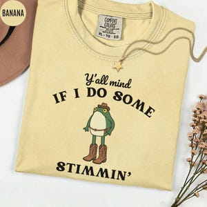 May include: A light yellow t-shirt with the text "Y'all mind if I do some stimmin'" and a cartoon frog wearing a cowboy hat and boots. A gold necklace with a star pendant is on the shirt. The shirt has a Comfort Colors tag.