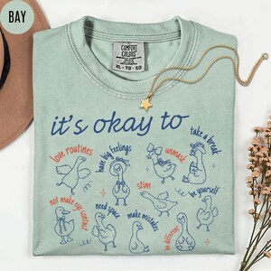 May include: A light green Comfort Colors t-shirt with the text "it's okay to" and various phrases and illustrations of ducks. A gold star necklace is draped across the shirt.
