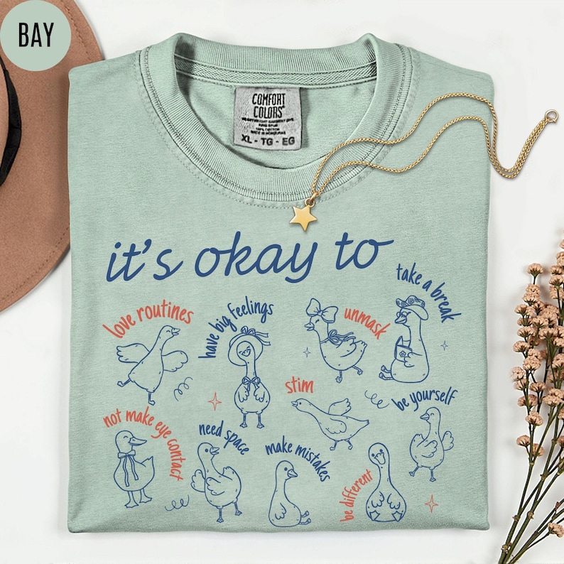 May include: A light green Comfort Colors t-shirt with the text "it's okay to" and various phrases and illustrations of ducks. A gold star necklace is draped across the shirt.
