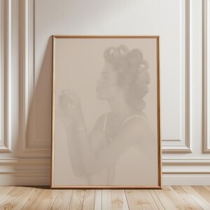 Op de afbeelding: Een ingelijste sepia-kleurige prent met een vrouw in profiel met haar haar in rollers. Ze draagt een tanktop en houdt iets vast. Het kunstwerk wordt tentoongesteld tegen een witte muur met decoratieve lijstwerk.