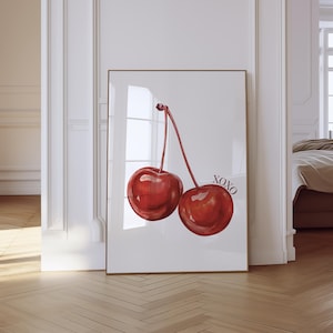 May include: A framed art print featuring a watercolor illustration of two ripe red cherries with long stems against a white background. The print includes the text "XOXO" in black. The frame is gold.
