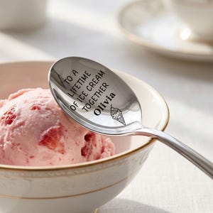 May include: A polished silver-toned metal spoon resting in a bowl of strawberry ice cream. The bowl of the spoon is engraved with the text TO A LIFETIME OF ICE CREAM TOGETHER Olivia and features a small graphic illustration of an ice cream cone.