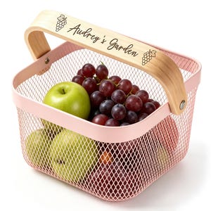 May include: A light pink metal mesh basket with a wooden handle engraved with the text Audrey's Garden and grape illustrations. The basket is filled with green apples and dark purple grapes, showcasing a functional storage design.
