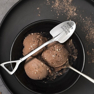 May include: A stainless steel ice cream shovel with the text "Olivia's Ice Cream Shovel" is placed in a black bowl filled with chocolate ice cream. The shovel has a spade-like scoop and a long handle. A second spoon is also in the bowl.