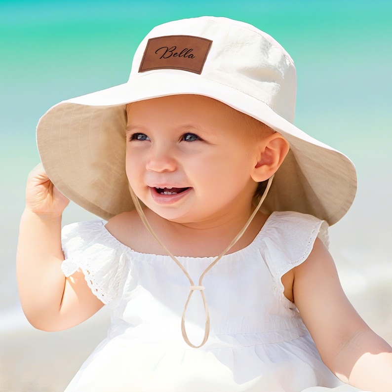 May include: A cream-colored sun hat with a wide brim and a brown leather patch that reads "Bella". The hat has a chin strap and is worn by a smiling child. The background is a blurred beach scene.