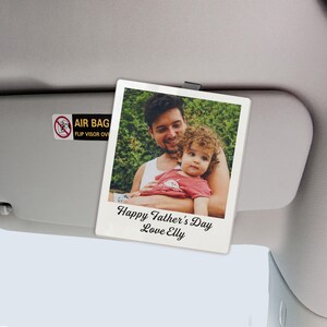 May include: A white rectangular car visor photo featuring a smiling man holding a child. The photo has a white border and the text "Happy Father's Day Love Elly" in black script. The visor is attached to a car's interior.